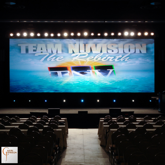 Team Nu Vision Conference with a large rear projection screen up stage with a row of white lights shining downstage for back light.