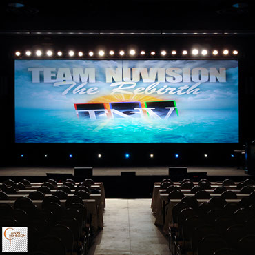 Team Nu Vision Conference with a large rear projection screen up stage with a row of white lights shining downstage for back light.