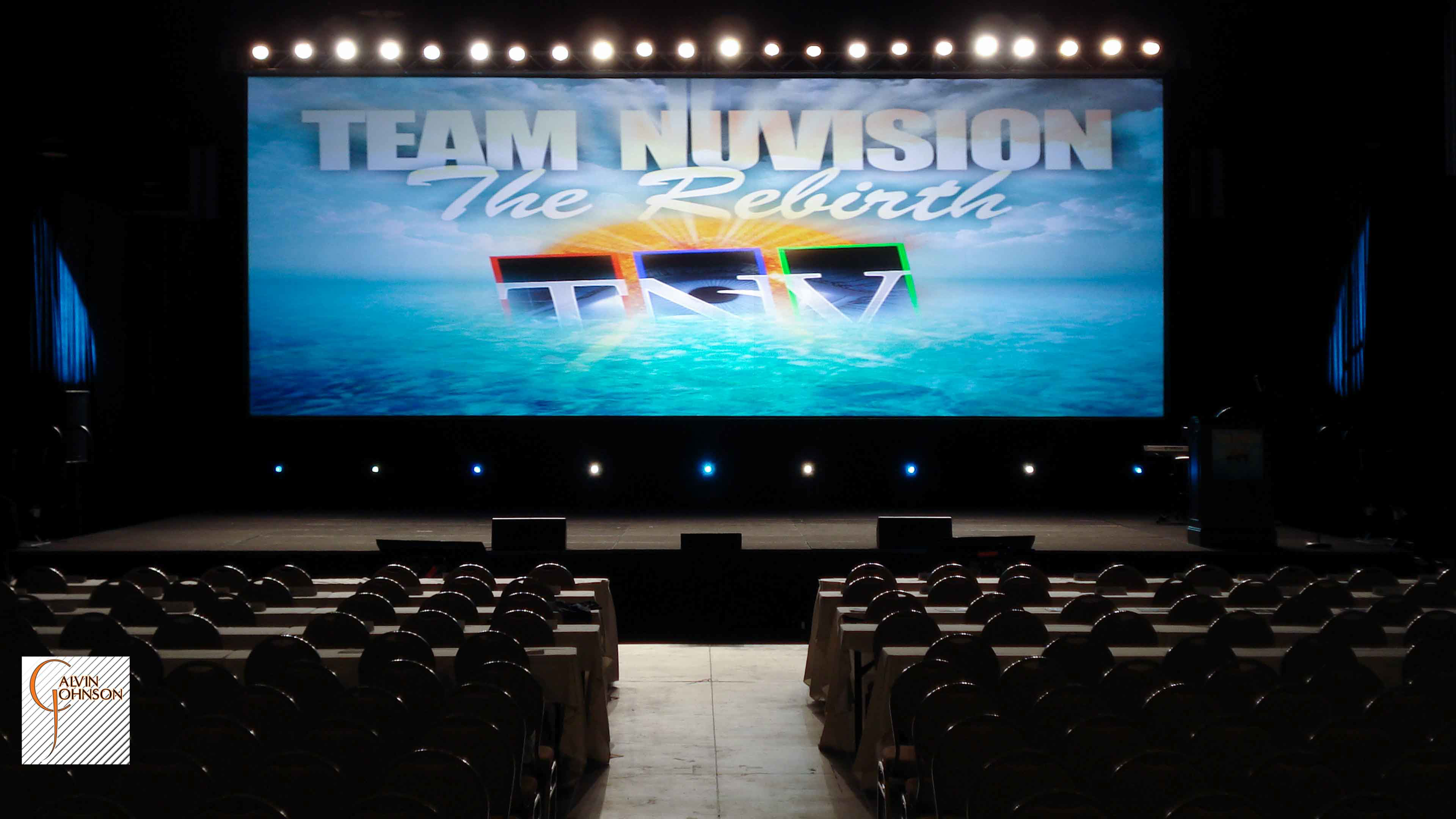 Team Nu Vision Conference with a large rear projection screen up stage with a row of white lights shining downstage for back light.